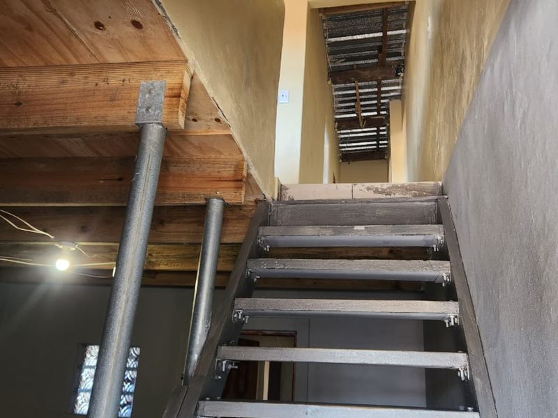 An unfinished interior space featuring a metal staircase, exposed timber joists, and steel support columns, currently in a shell condition requiring completion.