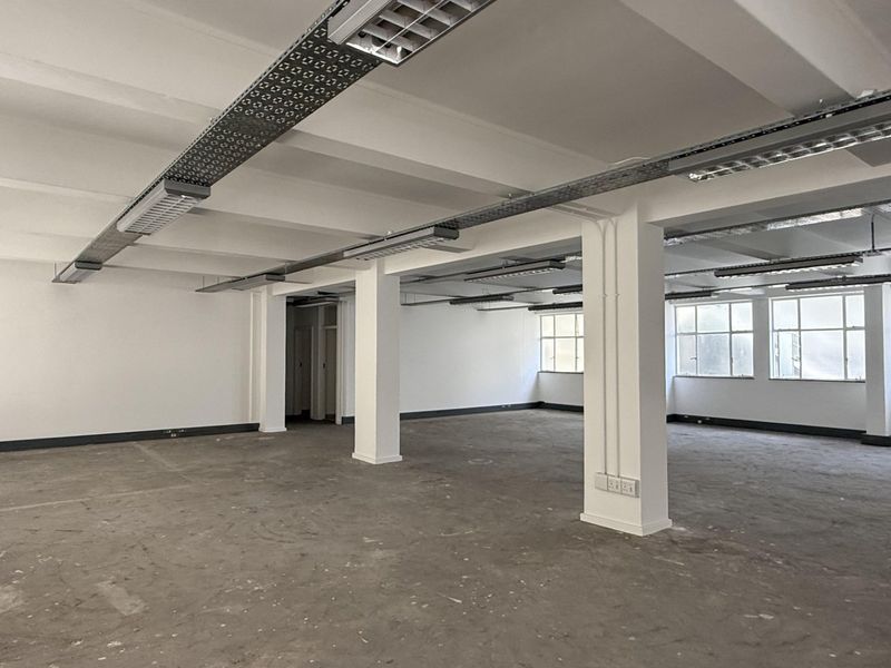 Spacious open-plan office area featuring exposed ceiling beams, industrial cable trays, large windows, and durable concrete flooring, offering a versatile blank canvas workspace.