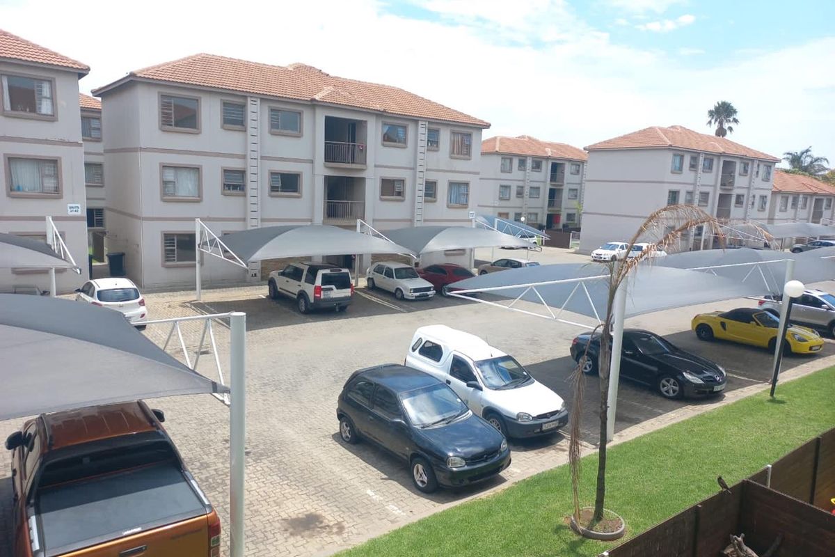 An elevated view of an apartment complex parking area featuring paved bays with shade cloth carports and surrounding landscaped lawn areas.
