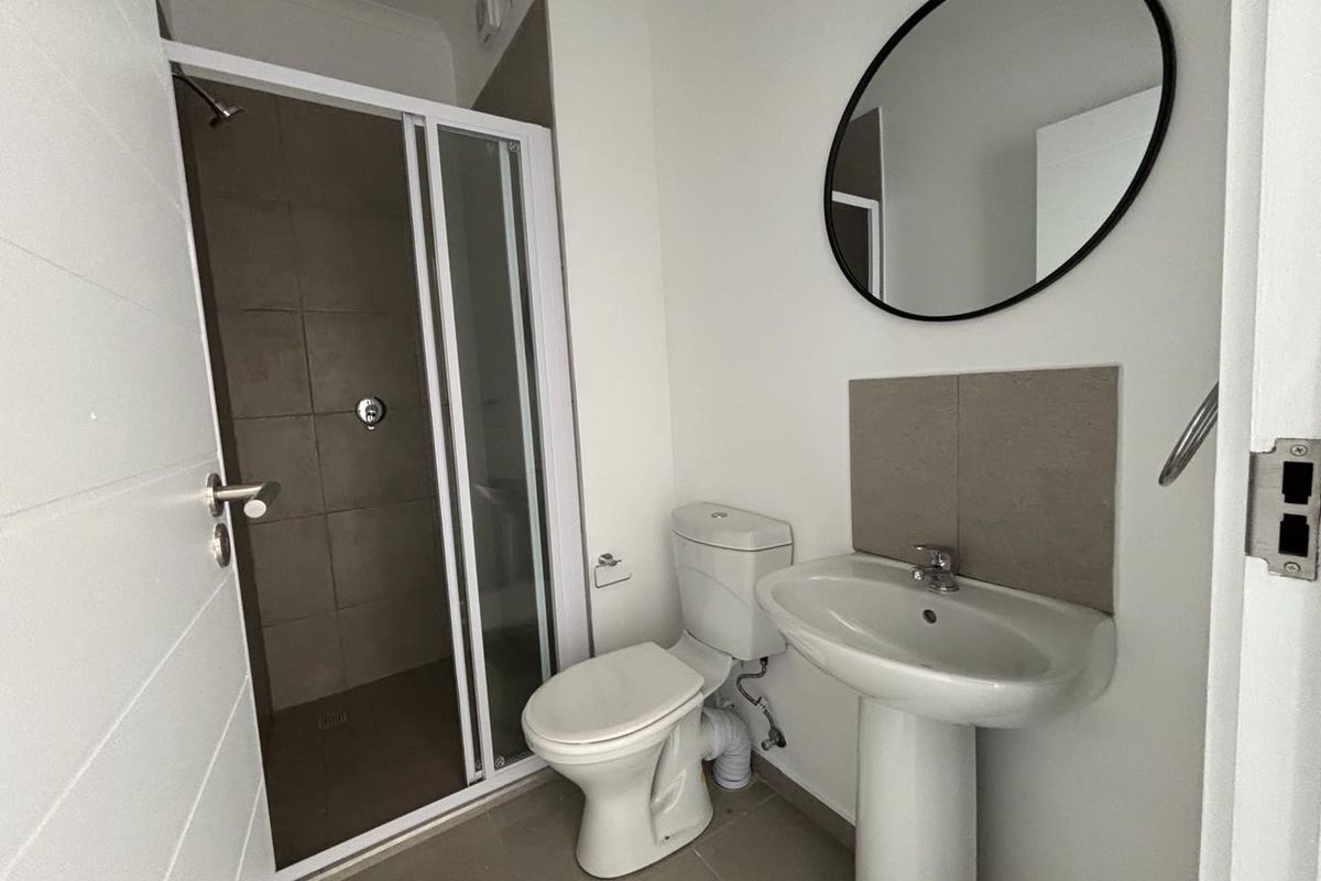 A clean and functional bathroom featuring a glass-enclosed shower, pedestal basin, and toilet with neutral wall tiling.