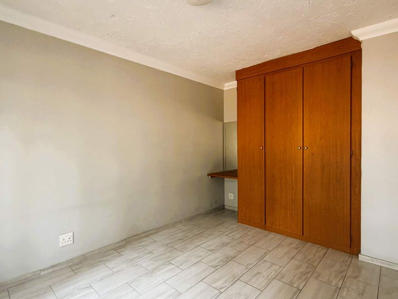 A bright, empty bedroom featuring light-toned tiled flooring, a built-in wooden wardrobe, and an integrated vanity desk.
