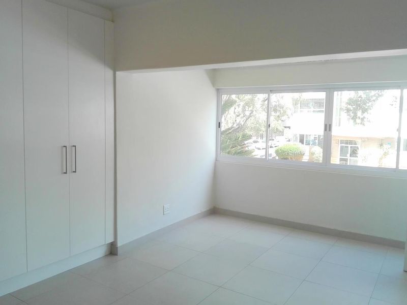 A bright, empty bedroom featuring neutral tiled flooring, built-in white cupboards, and large windows providing natural light.