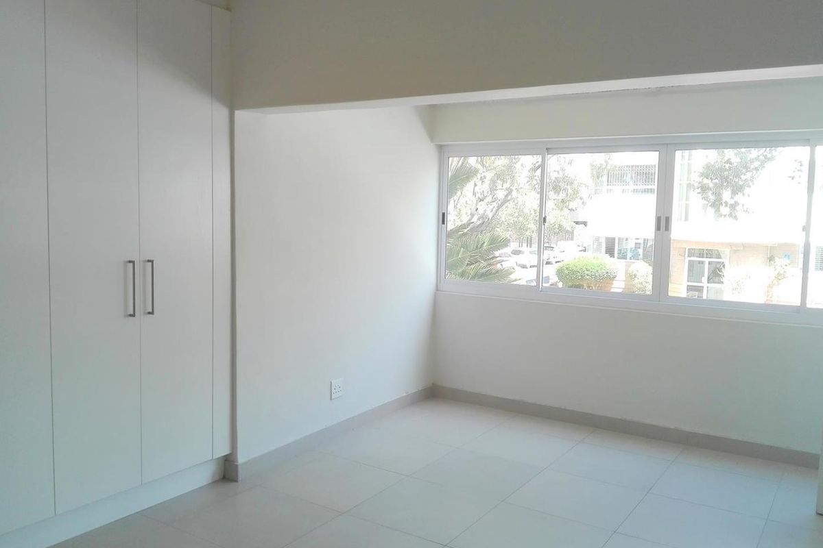 A bright, empty bedroom featuring neutral tiled flooring, built-in white cupboards, and large windows providing natural light.