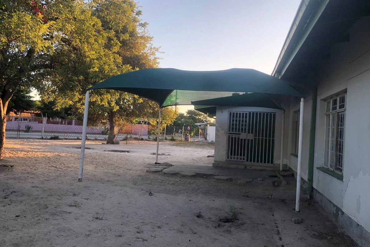 Rear exterior area featuring a shade sail structure, security gate, and metal window frames, set against a sandy yard with visible exterior wall wear.