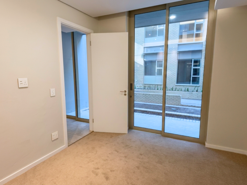 A bright, empty bedroom featuring neutral carpeting, a sliding glass door leading to an outdoor area, and a doorway connecting to an adjacent space.