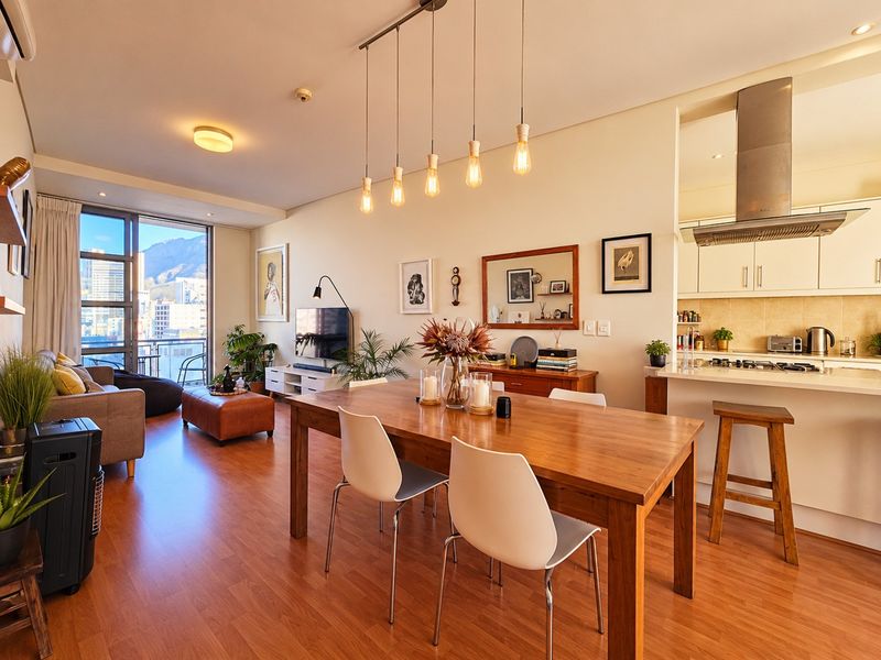 A bright open-plan living, dining, and kitchen area featuring laminate flooring, modern cabinetry, stone countertops, and access to a balcony with city views.