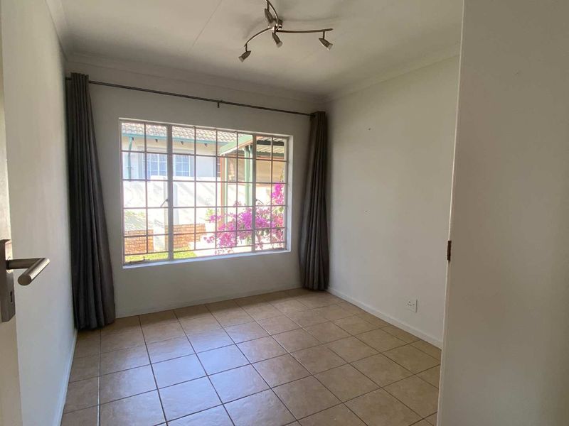 A bright, empty bedroom featuring neutral tiled flooring, a ceiling-mounted light fixture, and a large window with security bars overlooking the exterior.