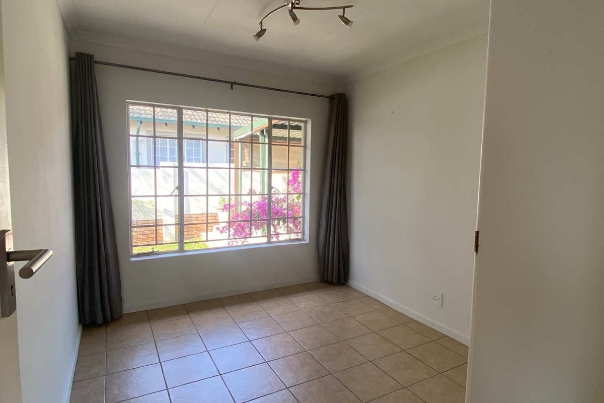 A bright, empty bedroom featuring neutral tiled flooring, a ceiling-mounted light fixture, and a large window with security bars overlooking the exterior.