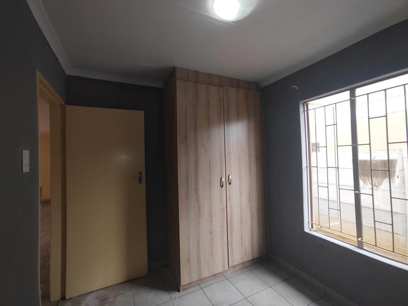 A clean, empty bedroom featuring tiled flooring, a built-in wooden wardrobe, and a window fitted with security bars.