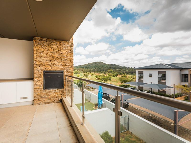 A spacious covered balcony featuring a built-in braai, stone cladding, and a glass balustrade, offering scenic views of the surrounding landscape.