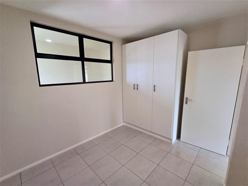 A clean, modern bedroom featuring neutral tiled flooring, a built-in white wardrobe, and an internal window providing natural light flow.