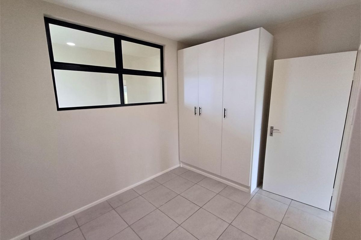 A clean, modern bedroom featuring neutral tiled flooring, a built-in white wardrobe, and an internal window providing natural light flow.