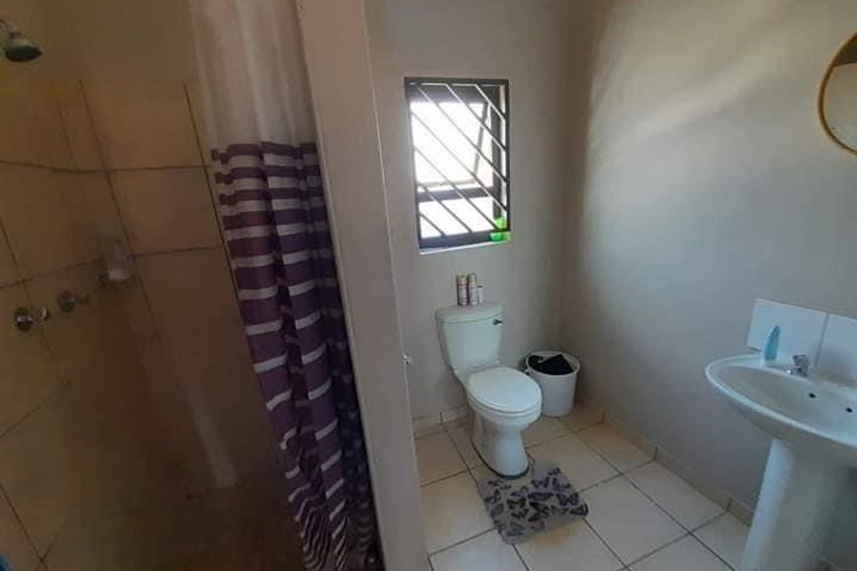 A functional bathroom featuring a shower with curtain, pedestal basin, and toilet, complemented by a security-grilled window and tiled flooring.