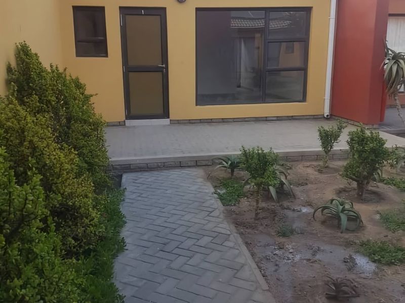 Rear exterior view of the apartment featuring a paved walkway, small patio area, and established garden beds with aluminium window and door frames.