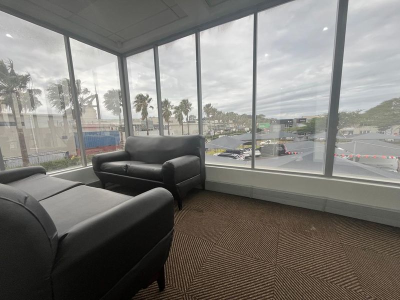 A bright office space featuring extensive floor-to-ceiling windows providing natural light and exterior views, finished with carpeted flooring and a suspended ceiling.