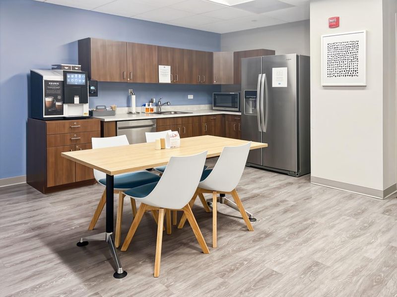 A modern office kitchenette featuring wood-tone cabinetry, stainless steel appliances, a sink, and durable laminate flooring, providing a functional break area for staff.