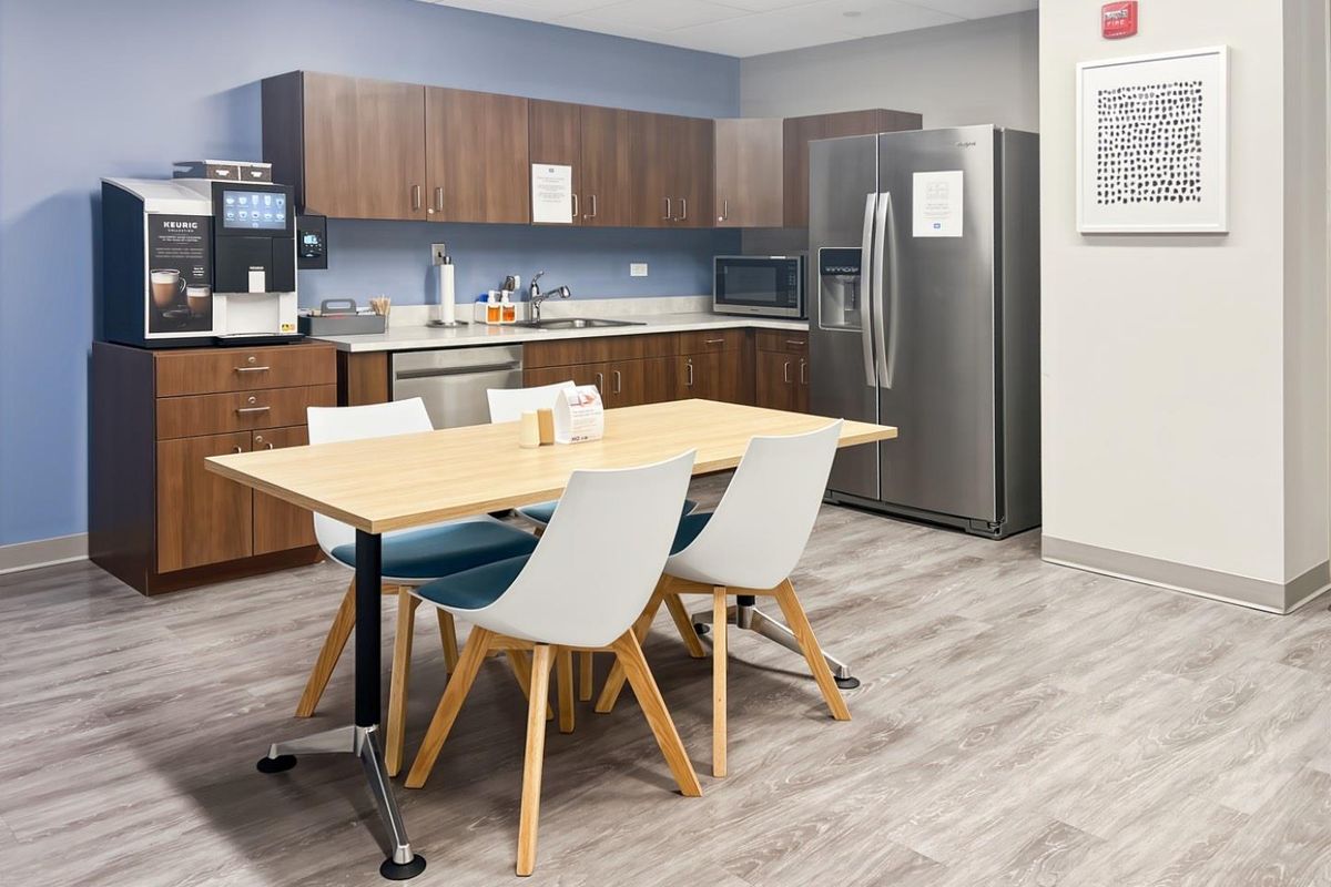 A modern office kitchenette featuring wood-tone cabinetry, stainless steel appliances, a sink, and durable laminate flooring, providing a functional break area for staff.