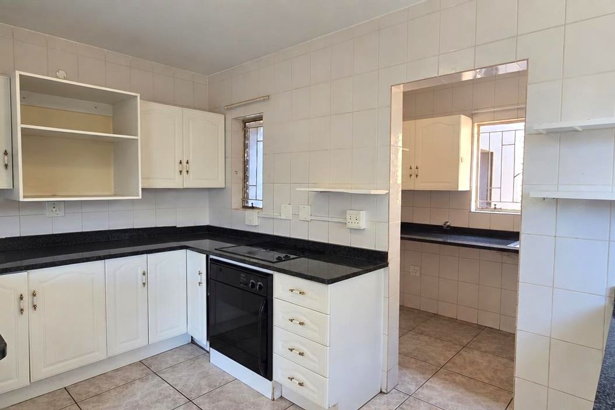 A functional kitchen featuring white cabinetry, dark granite countertops, built-in oven and hob, with an adjoining scullery area and tiled finishes throughout.