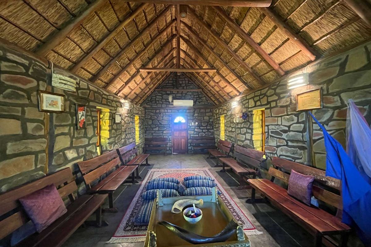 A rustic interior space featuring exposed stone walls, a high vaulted thatched roof with timber beams, and built-in wooden bench seating.