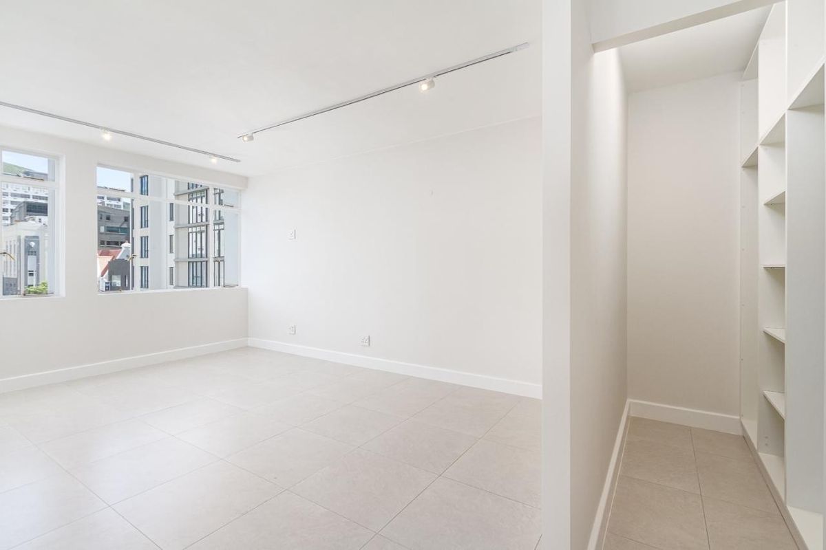 A bright, empty bedroom featuring neutral tiled flooring, built-in shelving, and modern track lighting with large windows offering an urban view.
