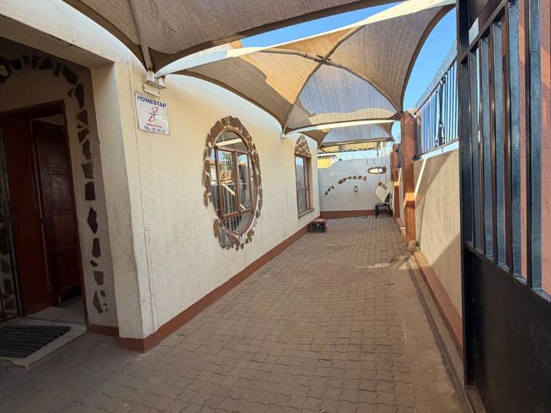 A paved outdoor walkway featuring a large shade sail, decorative stone window detailing, and secure metal gate access.