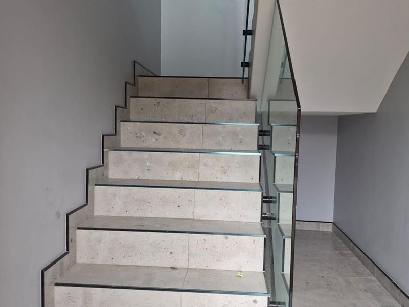 A modern interior staircase featuring stone-look tiled steps, a sleek glass balustrade, and a minimalist metal handrail.