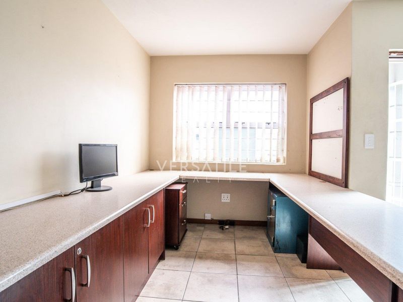 Functional office space featuring extensive built-in desk workspace, laminate cabinetry for storage, tiled flooring, and a window fitted with vertical blinds and security bars.