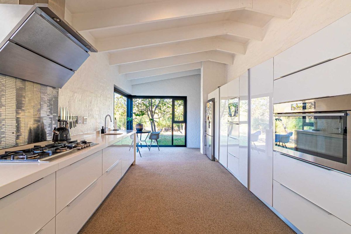 Modern galley kitchen featuring sleek white cabinetry, integrated stainless steel appliances, gas hob, extractor fan, and exposed beam vaulted ceilings with garden views.