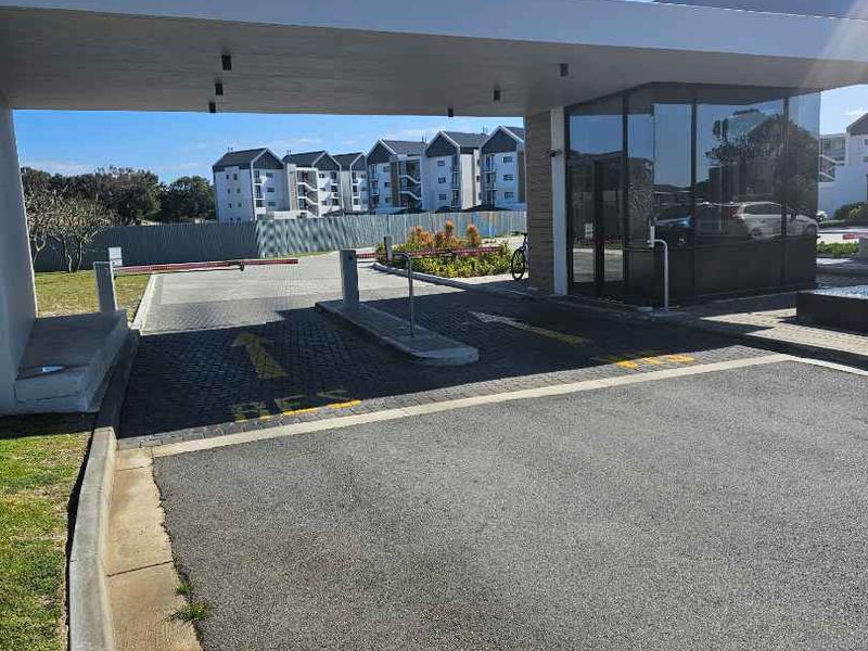 Modern security gatehouse entrance with a boom gate, paved driveway, and glass-fronted guard office, providing controlled access to the apartment complex.