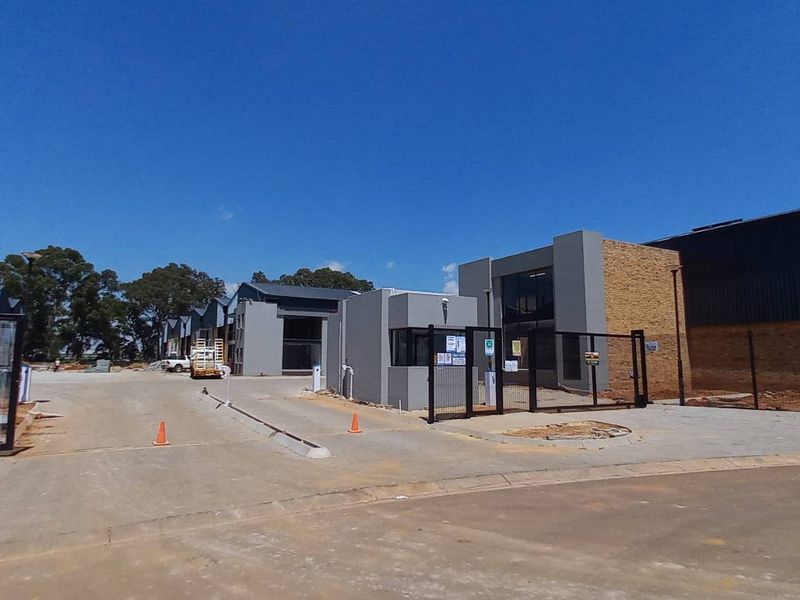 Front view of an industrial park entrance featuring a security gatehouse, paved access roads, and modern warehouse units with metal roofing and brick finishes.