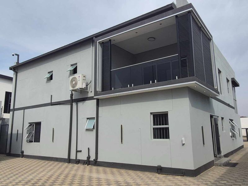 Modern two-story residential exterior featuring a private balcony with glass balustrades, paved driveway, and contemporary grey facade finishes.