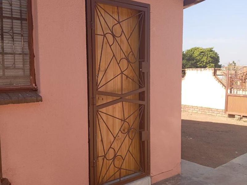 Exterior view of a rear building entrance featuring a wooden door with a security gate, window with burglar bars, and concrete apron.