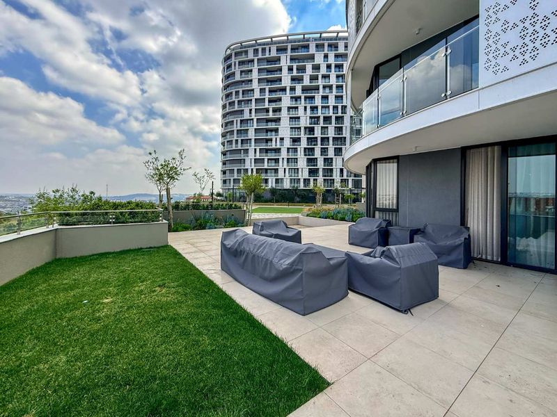 Spacious modern balcony featuring tiled flooring, glass balustrades, and direct access to a private lawn area with views of the surrounding apartment complex.