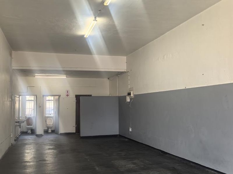 An empty, utilitarian commercial space featuring a concrete floor, partition wall, and rear access to two small toilet cubicles with wash basin.