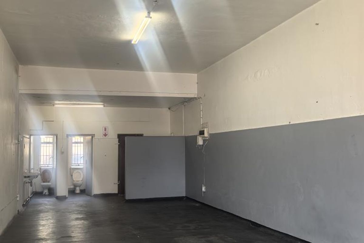 An empty, utilitarian commercial space featuring a concrete floor, partition wall, and rear access to two small toilet cubicles with wash basin.