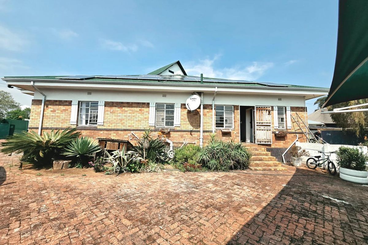 Rear view of a single-story brick house featuring a green metal roof with solar panels and a paved courtyard area.
