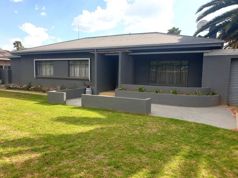 Front elevation of a single-story house featuring a grey painted facade, corrugated metal roof, and a paved driveway with integrated retaining walls.
