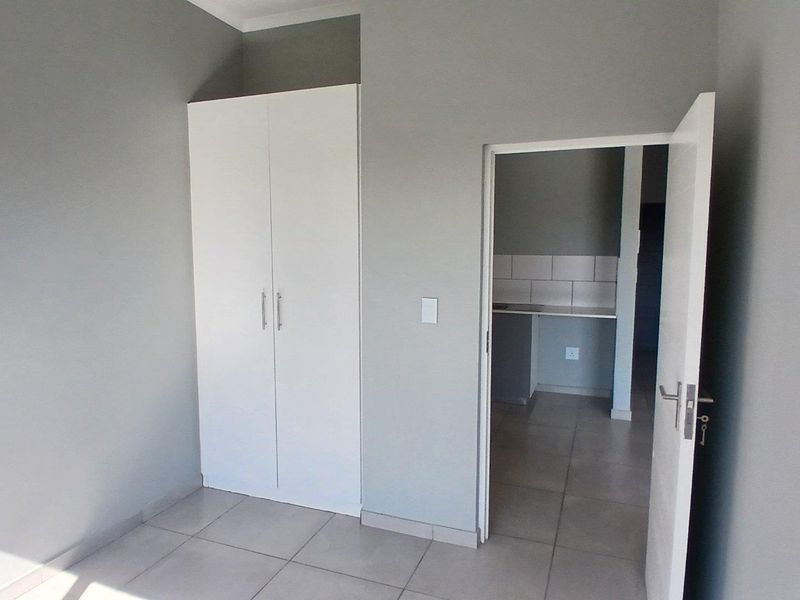 A clean, empty bedroom featuring neutral grey walls, tiled flooring, and a built-in white wardrobe, with a doorway leading to an adjacent kitchen area.