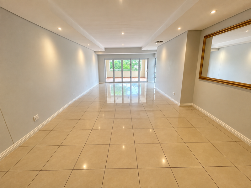 A spacious, bright, and empty living area featuring polished porcelain floor tiles, recessed downlighting, and large sliding glass doors leading to a balcony.
