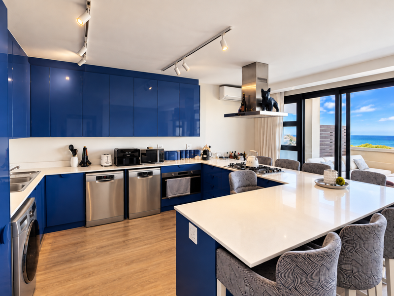 Modern kitchen featuring high-gloss blue cabinetry, quartz countertops, integrated appliances, and a breakfast bar, with direct access to a balcony and ocean views.