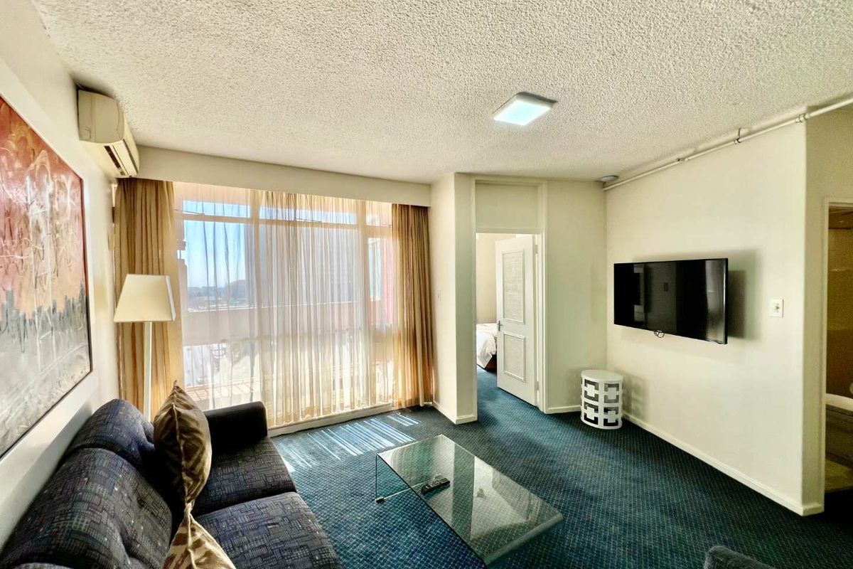 A living room featuring wall-mounted air conditioning, large windows with natural light, and access to an adjoining bedroom.