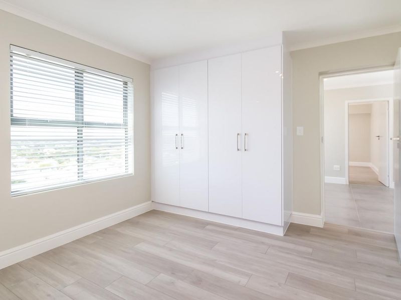 A bright, empty bedroom featuring modern white built-in cupboards, light-toned laminate flooring, and venetian blinds, offering a clean and neutral living space.