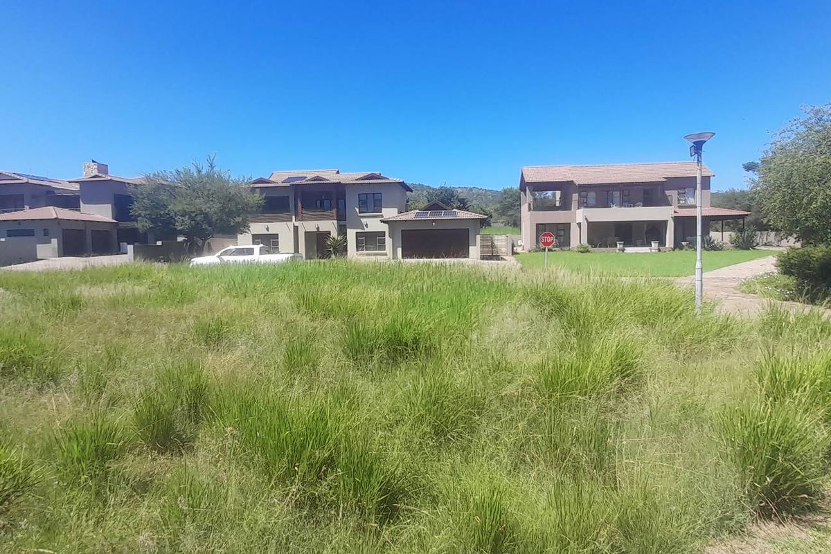A view of a vacant residential plot featuring tall grass, with established residential properties visible in the background.