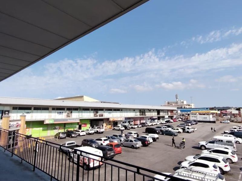 Elevated balcony area overlooking a commercial parking lot and retail storefronts, featuring a metal safety railing and covered overhead structure.