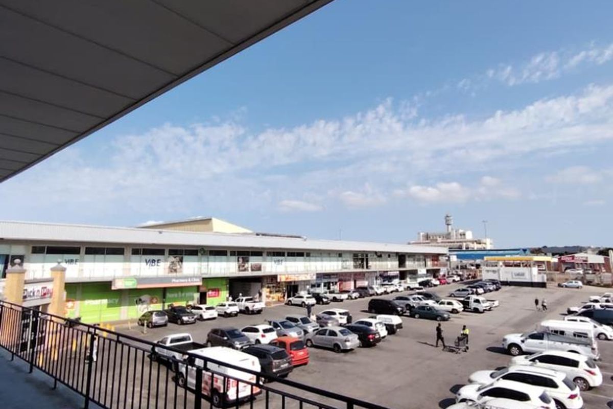 Elevated balcony area overlooking a commercial parking lot and retail storefronts, featuring a metal safety railing and covered overhead structure.