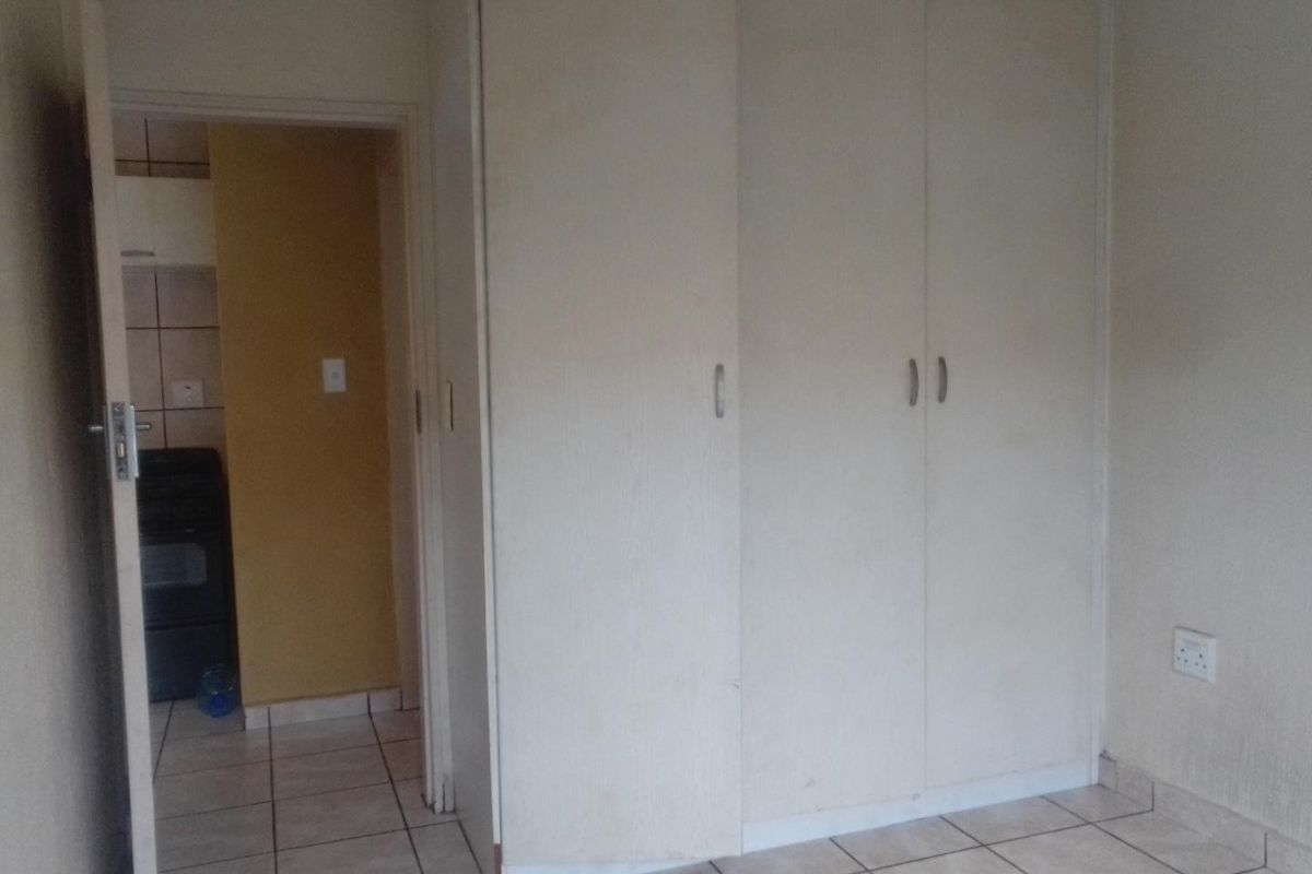 A bedroom featuring built-in cupboards and tiled flooring, with a view into an adjacent kitchen area.