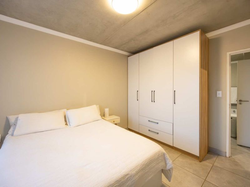 A modern bedroom featuring a large built-in wardrobe, neutral walls, and an exposed concrete ceiling, with an adjoining bathroom visible through the doorway.