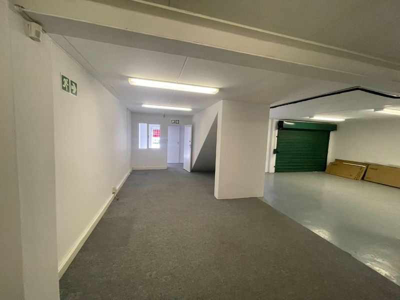 A spacious, vacant utility or storage area featuring carpeted and concrete flooring, fluorescent lighting, and a secure roller shutter door.
