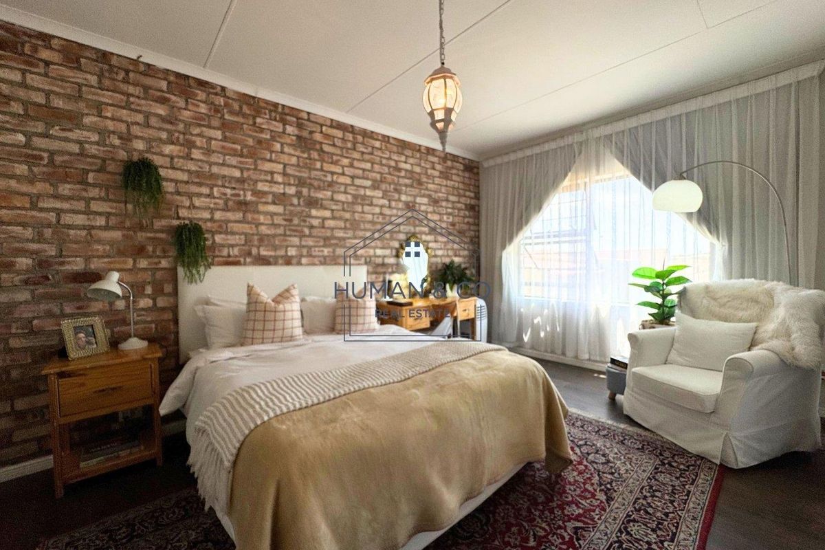 A cozy bedroom featuring a rustic exposed brick feature wall, large window with curtains, and dark wood-look flooring.
