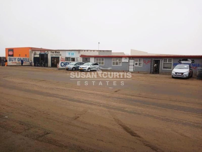 Front elevation of an industrial property featuring multiple commercial units with roller shutter doors and signage, situated along an unpaved road.